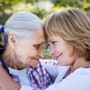 photo of an elderly woman on the left, wearing a plaid shirt with a white t-shirt underneath it touches her forehead to the younger woman on the right as they look into each other's eyes and smile.