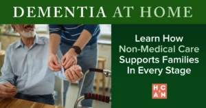 Seated older man smiling as a caregiver helps button his shirt cuff indoors, with a walker visible in the foreground, illustrating non-medical dementia care at home.