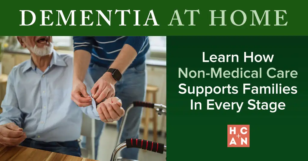 Seated older man smiling as a caregiver helps button his shirt cuff indoors, with a walker visible in the foreground, illustrating non-medical dementia care at home.